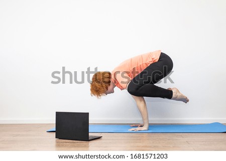 Yoga at home online. Quarantine. Coronavirus pandemic. Red-haired girl doing yoga asanas, looking at the laptop screen. Blue yoga mat. Against the background of a white wall.