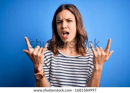 Young beautiful blonde girl wearing casual sweater standing over blue isolated background shouting with crazy expression doing rock symbol with hands up. Music star. Heavy concept.