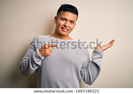 Young handsome latin man wearing casual sweater standing over isolated white background amazed and smiling to the camera while presenting with hand and pointing with finger.