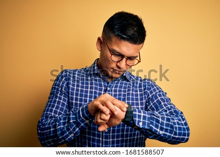 Young handsome latin man wearing casual shirt and glasses over yellow background Checking the time on wrist watch, relaxed and confident