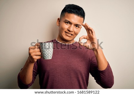 Young handsome latin man drinking cup of coffee over isolated white background doing ok sign with fingers, excellent symbol