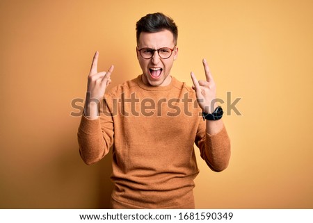 Young handsome caucasian man wearing glasses and casual winter sweater over yellow background shouting with crazy expression doing rock symbol with hands up. Music star. Heavy concept.