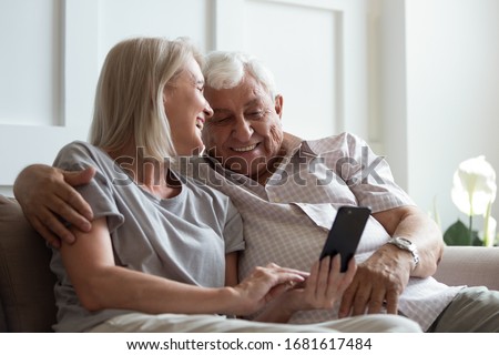 Happy smiling older woman and man hugging, laughing mature wife and husband cuddling, using phone together, having fun, watching funny video or chatting online, sitting on cozy sofa at home Royalty-Free Stock Photo #1681617484