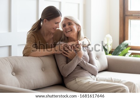 Loving adult daughter hugging older mother, standing behind couch at home, family enjoying tender moment together, young woman and mature mum or grandmother looking at each other, two generations Royalty-Free Stock Photo #1681617646