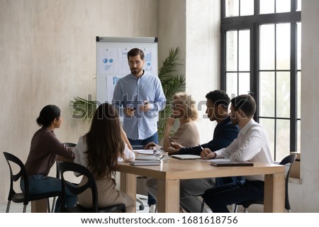 Confident male Caucasian team leader head meeting in office, present project on whiteboard, man speaker or coach talk make flip chart presentation to multiracial colleagues at briefing Royalty-Free Stock Photo #1681618756