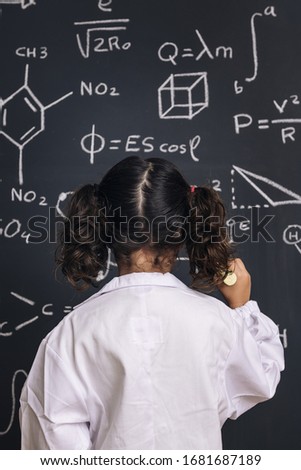 rear view of a little girl science student in lab coat writing on school blackboard with hand drawings science formula pattern, back to school and successful female career concept, vertical photo