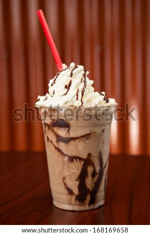 Chocolate milkshake in a plastic cup, topped with whipped cream and a red straw.  It is on a wood table with a corrugated copper wall in the background. Royalty-Free Stock Photo #168169658