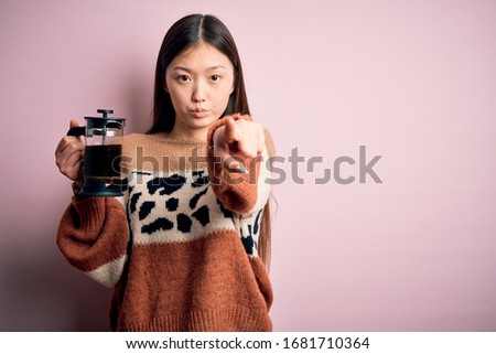 Young asian woman making a glass of coffe using french press coffee maker over pink background pointing with finger to the camera and to you, hand sign, positive and confident gesture from the front
