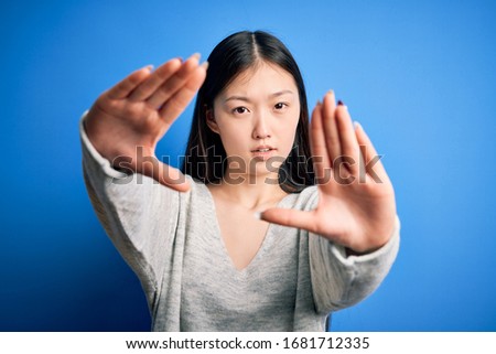 Young beautiful asian woman wearing casual sweater standing over blue isolated background doing frame using hands palms and fingers, camera perspective