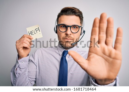 Young call center operator business man with blue eyes offering support holding paper note with open hand doing stop sign with serious and confident expression, defense gesture