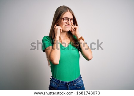 Young beautiful redhead woman wearing casual green t-shirt and glasses over white background covering ears with fingers with annoyed expression for the noise of loud music. Deaf concept.