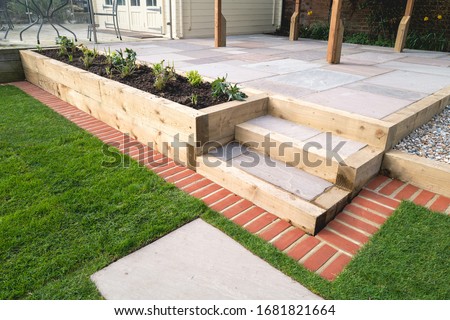 New steps in a garden or back yard leading to a raised patio, alongside a new raised flowerbed made using wooden sleepers. A mowing strip of bricks is in front of newly laid turf.  Royalty-Free Stock Photo #1681821664