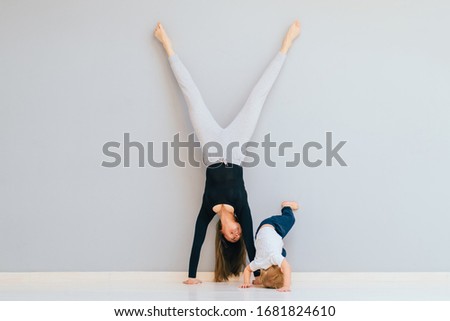 Sport and yoga. Strong young athletic woman doing exercise her little son try doing the same. Handstand against grey wall, full length. Conception of motherhood youth and energy.