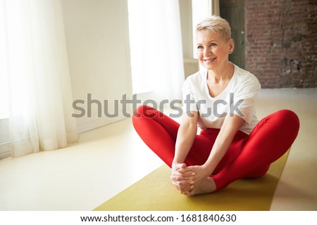 Joyful athletic Caucasian female sitting on yoga mat with knees bent, holding hands on feet, doing hips opening poses, smiling, enjoying stretching exercises. Flexibility, stretching and pilates