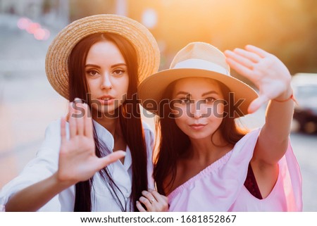 Two pretty girls take selfie in the summer outdoors at sunset. Girlfriends have fun, laugh, smile and take pictures. Closeup romantic portrait of two young women in dresses and straw hats.
