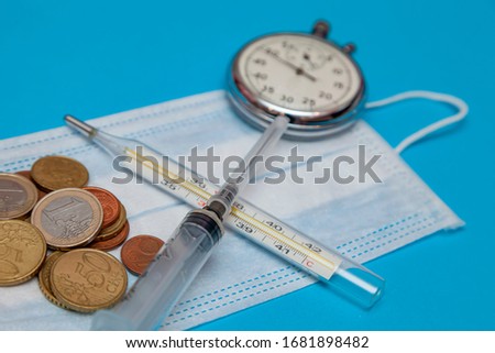 White medical mask lies on a blue background. Also lie on the mask money, coins of one euro, 50 euro cent.Next to the coins are a silver watch and a disposable syringe with a thermometer.