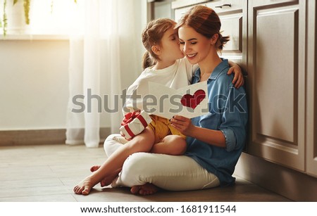 Happy little girl congratulating smiling mother and giving card with red heart during holiday celebration at home
