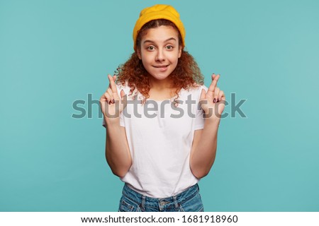 Hoping young lovely curly brunette woman dressed in casual clothes biting worringly underlip and crossing her fingers for good luck while making wish, isolated over blue background