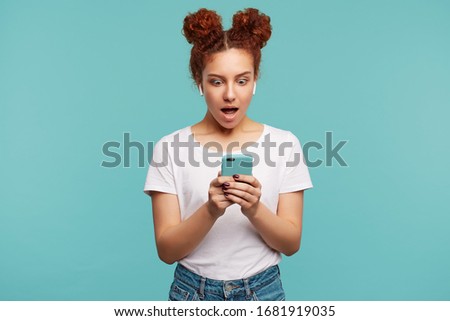 Open-eyed young curly brunette female looking amazedly at screen of her smartphone while reading shocking news, dressed in casual clothes while posing over blue background