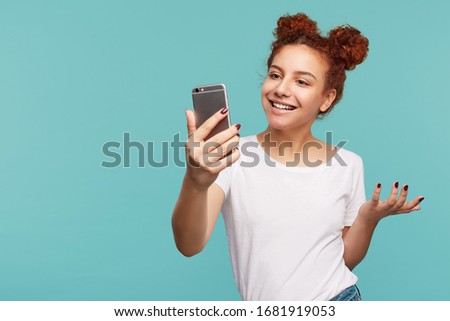 Indoor shot of positive young curly brunette female with bun hairstyle keeping mobile phone in raised hand while having video chat with her friend, isolated over blue background