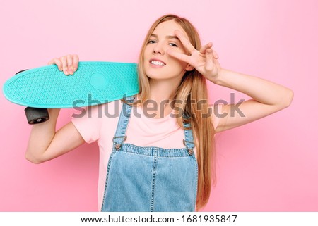 Portrait of an attractive skater girl holding a skateboard and showing a peace sign isolated on a pink background in a Studio