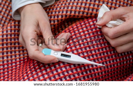 Woman sitting on sofa covered with colorful blanket, taking her temperature. Thermometer showing 38,2 degrees on Celsius scale (100,8 F). Close up.