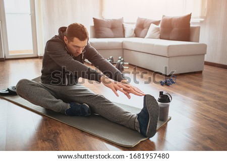 Young ordinary man go in for sport at home. Freshman or beginner stretching with tow hands to one leg. Excercising alone in empty apartment. Try to keep fit and stay healthy with good body Royalty-Free Stock Photo #1681957480
