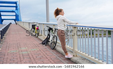 Curly-haired blonde girl on a bicycle trip, happy resting on a footpath on top of a bridge. Bike ride on the bike path over the bridge over the river. emotions joy, fun. inspire world
