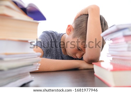 Tired disappointed schoolboy lowering his head sitting among pile of books, textbooks, school exercise books on his desk at home Royalty-Free Stock Photo #1681977946