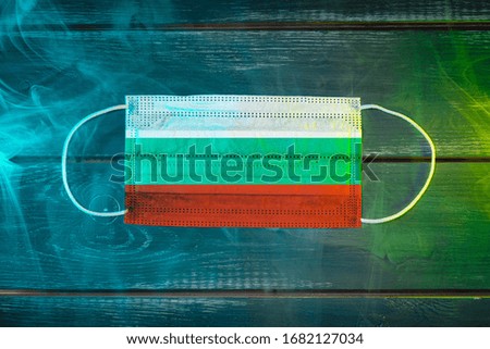 Medical mask for protection against airborne diseases, painted in the national flag of Bulgaria on a black background in blue-green smoke. Medical protection against airborne diseases, coronavirus