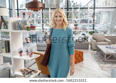 All for comfort. Happy woman in a blue dress walking through the furniture salon, where there are many things for the interior, smiling.