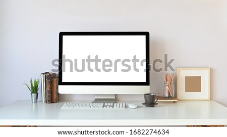 Photo of Computer monitor with white blank screen, stack of books, potted plant, wireless keyboard, mouse, coffee cup, pencil holder and picture frame putting together on white working desk.