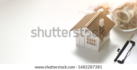 Photo of house model, clipboard, saving money in vase putting together on white table. Planning for buying a house concept.