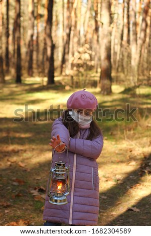 little beautiful girl in a protective mask from the crown of the virus stands in the forest and holds a kerosene lamp in which fire is burning