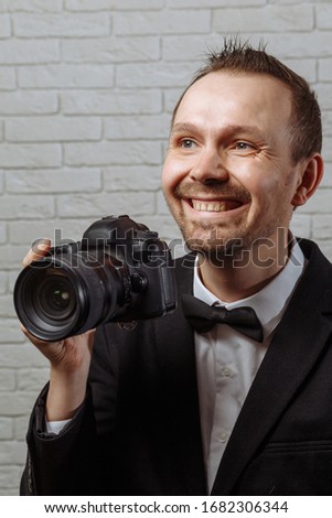 Young handsome male photographer with a beautiful smile in a black jacket, white shirt and black bow-tie holds a camera in his hand and smiles.
