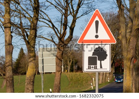 A red and white warning sign and a graphic representation with a truck and a tree warns of a possible collision with trees or tree tops. In the background are trees and a blue car.