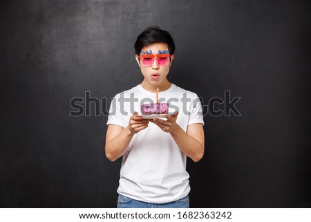 Birthday, celebration and party concept. Portrait of young asian guy grew up, wear funny sunglasses celebrating b-day blowing out candle on cake to make wish, standing black background