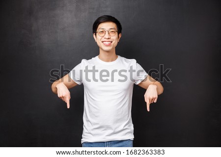 Studio portrait of happy handsome asian male student in white t-shirt, inviting new members join his team start career in business, pointing fingers down look here sign, smiling camera