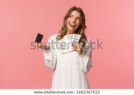 Portrait of pleased attractive blond girl in white dress, daydreaming about what buy, shopping with girlfriends, holding money and credit card, use cash, standing pink background