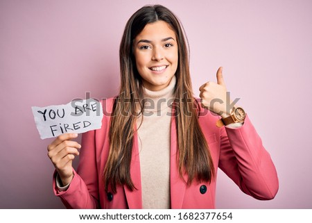 Young beautiful girl holding paper with you are fired message over isolated pink background happy with big smile doing ok sign, thumb up with fingers, excellent sign