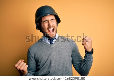 Young handsome businessman wearing military helmet over isolated yellow background very happy and excited doing winner gesture with arms raised, smiling and screaming for success. Celebration concept.