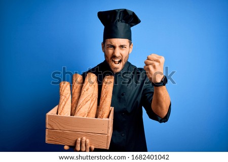 Young cooker man with beard wearing uniform holding box with bread over blue background annoyed and frustrated shouting with anger, crazy and yelling with raised hand, anger concept