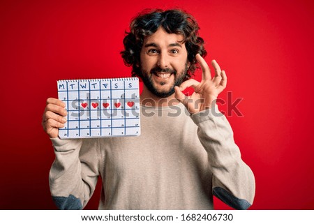 Young handsome man with beard holding calendar over isolated red background doing ok sign with fingers, excellent symbol