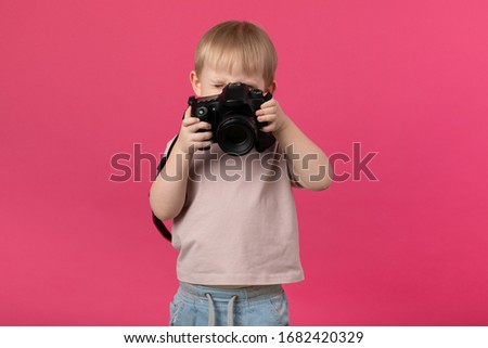 A kid boy of European appearance blond photographs with a camera. Close-up studio shot on a pink background for articles about recruiting for schools and courses, shooting training.
