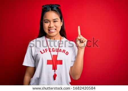 Young asian lifeguard girl wearing t-shirt with red cross using whistle over isolated background with a big smile on face, pointing with hand finger to the side looking at the camera.
