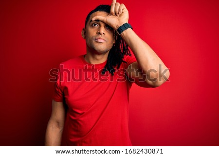 Young handsome african american afro man with dreadlocks wearing red casual t-shirt making fun of people with fingers on forehead doing loser gesture mocking and insulting.