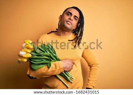 Young african american afro romantic man with dreadlocks holding bouquet of yellow tulips looking sleepy and tired, exhausted for fatigue and hangover, lazy eyes in the morning.