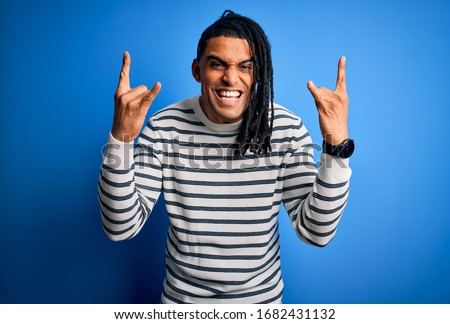 Young handsome african american afro man with dreadlocks wearing casual striped sweater shouting with crazy expression doing rock symbol with hands up. Music star. Heavy music concept.