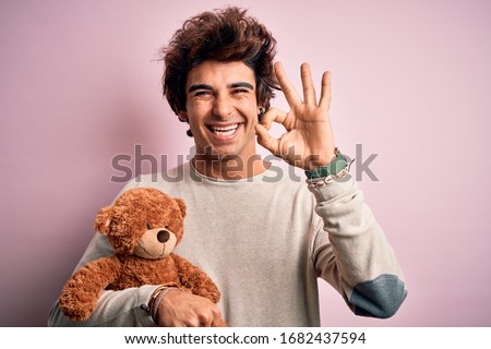 Young handsome man holding teddy bear standing over isolated pink background doing ok sign with fingers, excellent symbol