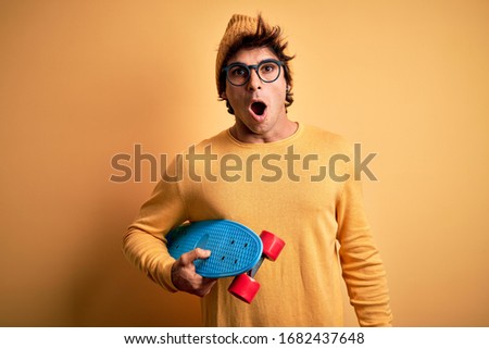 Young handsome student man holding skate wearing glasses over isolated yellow background scared in shock with a surprise face, afraid and excited with fear expression